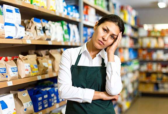 Une vendeuse fatiguée portant un tablier vert se tient la tête entre les rayons d'un magasins.